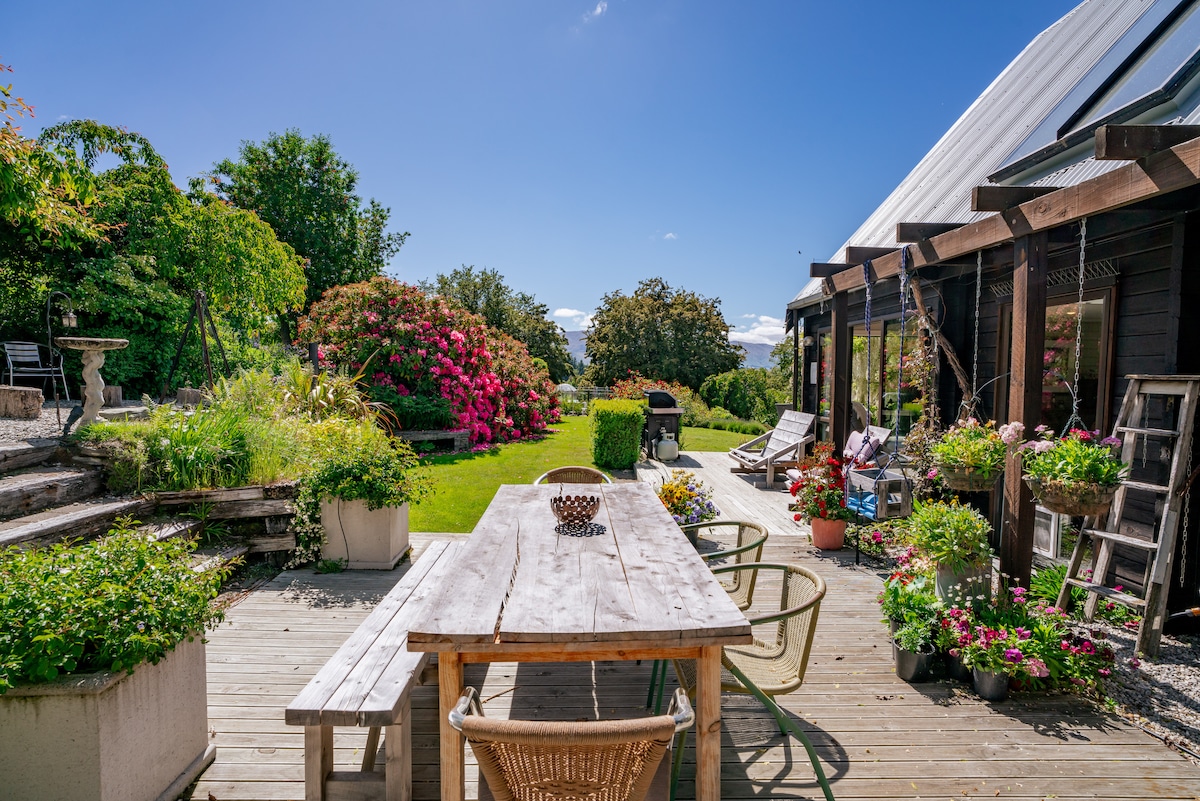 A spacious outdoor patio is depicted, featuring a wooden dining table surrounded by several chairs. Lush gardens with vibrant flowering plants and greenery enhance the serene environment. A barbeque area is visible, alongside pathways leading to a tranquil landscape under a clear blue sky.