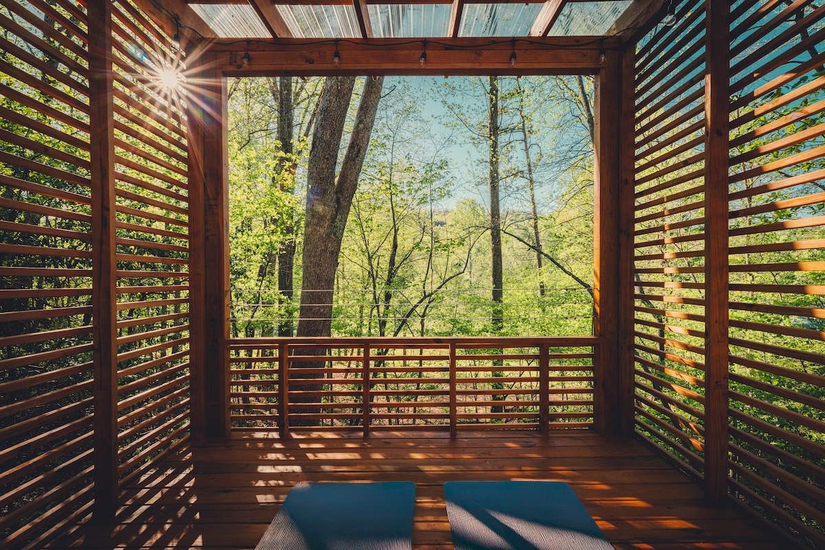 A serene observation deck is framed by wooden slats, allowing natural light to filter through. Two yoga mats are positioned on the warm wooden floor, while trees and vibrant greenery create a peaceful backdrop, providing a tranquil space for relaxation or exercise.