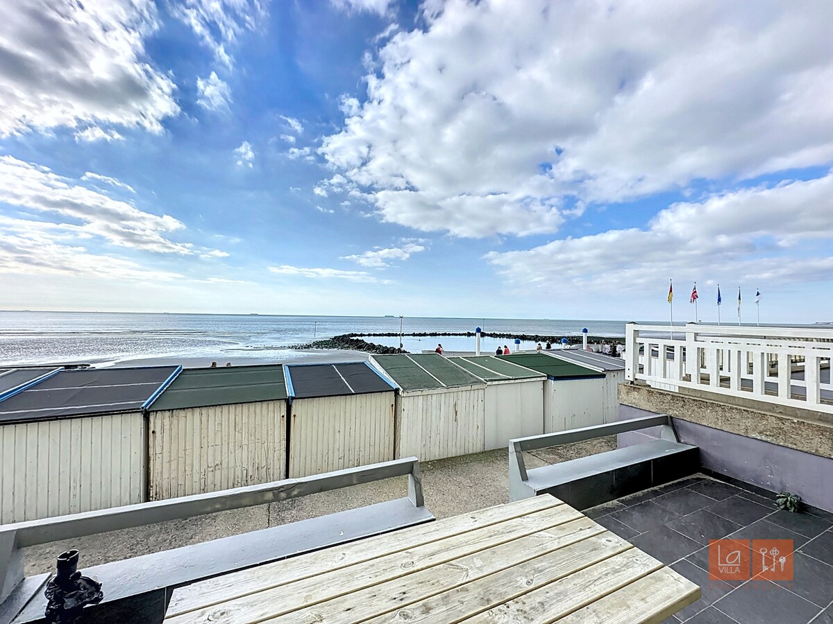 A terrace offers an expansive view of the beach and the sea, framed by a row of beach cabins. The sky features light clouds with hints of blue, while the horizon meets the water in the distance. A simple wooden table is positioned on the terrace.
