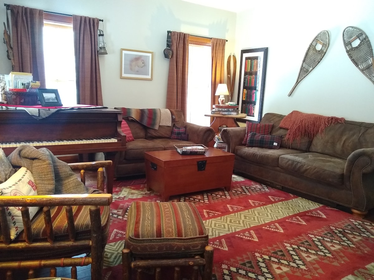 The living area features a mix of seating options, including a brown leather sofa and armchair paired with a wooden coffee table. A charming piano complements the cozy decor, which is enhanced by warm-toned curtains and a patterned area rug. Natural light filters through the windows, creating a welcoming ambiance.