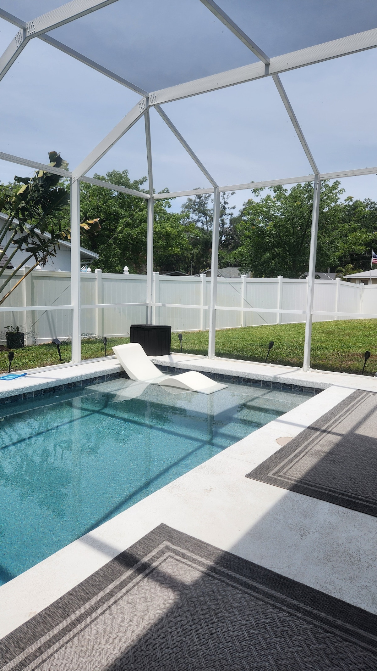A private pool area enclosed by a screen porch is displayed. The pool features a pair of loungers positioned on the water, with surrounding greenery visible through the frame. Concrete deck surrounds the pool, complemented by patterned outdoor rugs.
