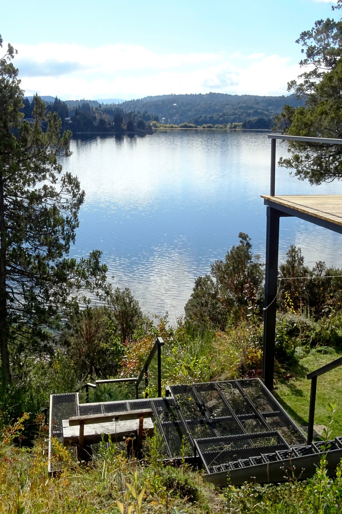A staircase leads down to a serene lake, surrounded by trees and lush greenery. The tranquil water reflects the sky and distant hills, creating a calming natural view. A small wooden bench is positioned beside the steps, offering a peaceful spot to relax.