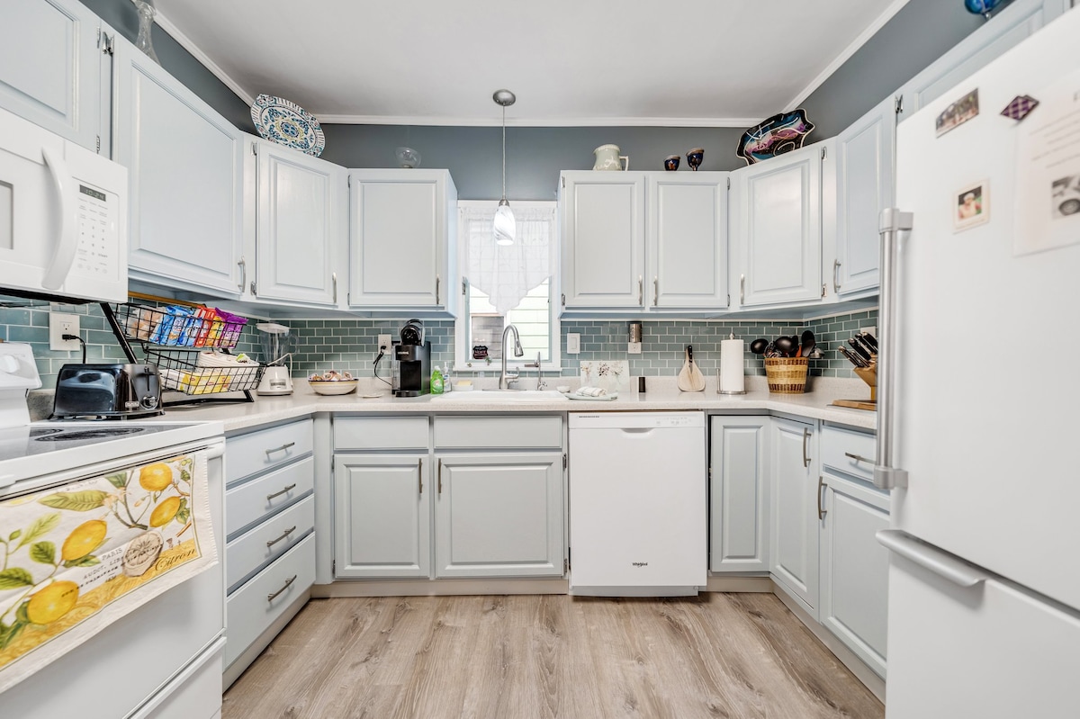 The kitchen features light blue cabinets and modern appliances, including a refrigerator and microwave. A white countertop holds a coffee maker and a basket. Tiled backsplash adds a subtle texture. Natural light enters through a window, enhancing the overall bright and functional space.