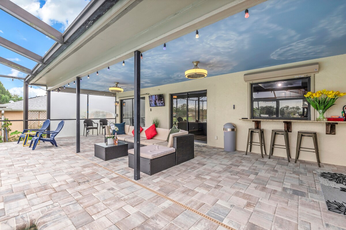 An outdoor lanai area features a spacious seating arrangement with modern furniture and decorative cushions. Soft blue sky imagery adorns the ceiling, while potted flowers add a fresh touch. A dining bar area is visible, complementing the inviting atmosphere for relaxation and entertainment.