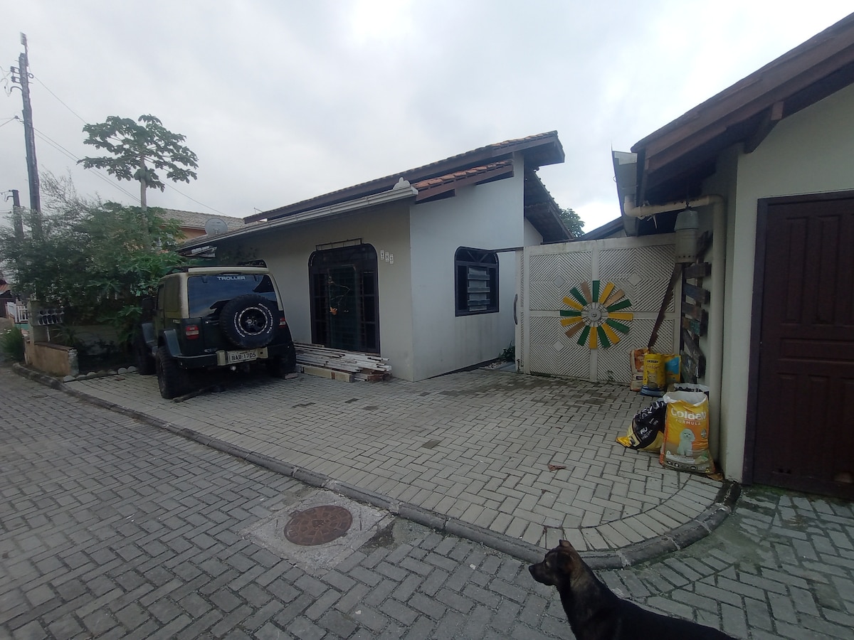A single-story building with a rustic facade is visible, accompanied by a paved driveway. A parked vehicle is positioned in front of the entrance, while nearby are decorative trees and a large wall mural featuring a sun design. A friendly dog stands on the driveway.