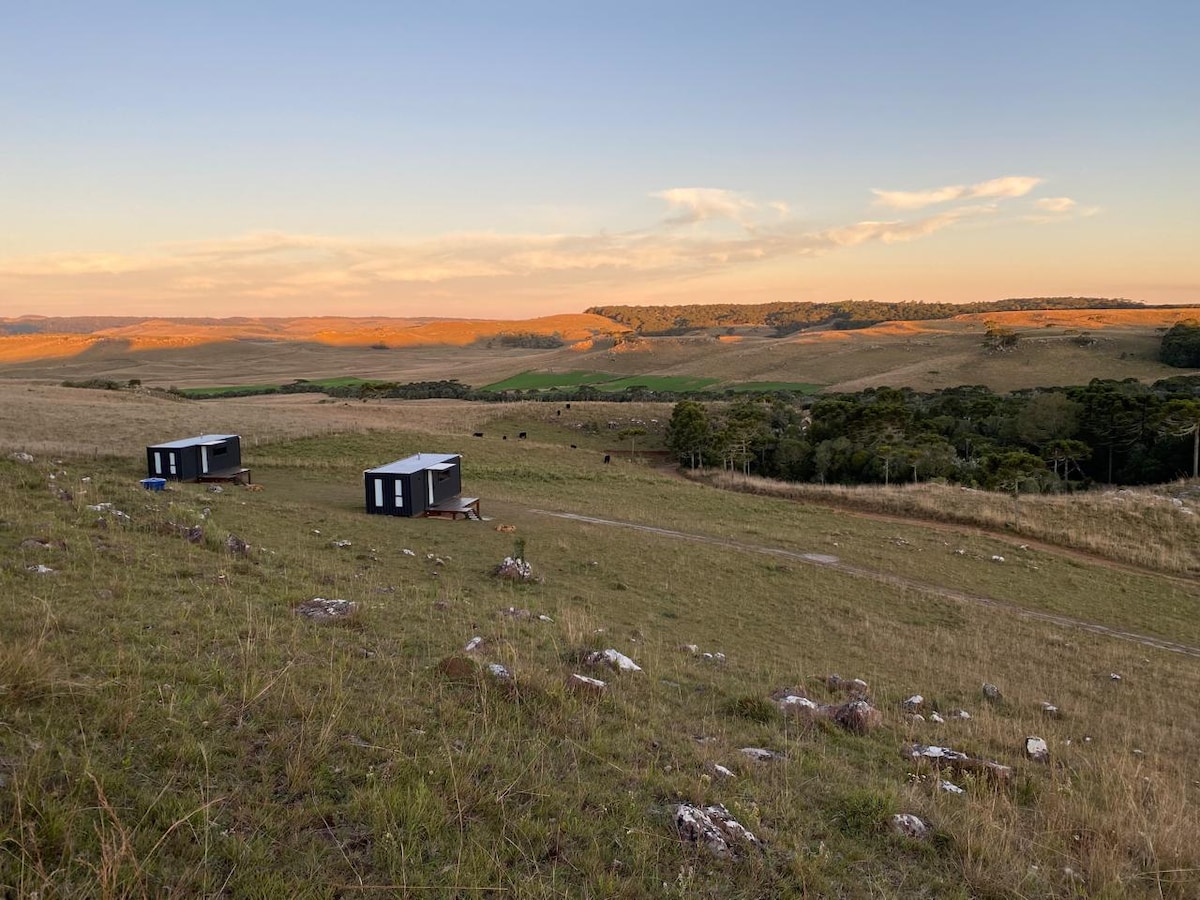Two modern studios are situated on a hillside surrounded by open fields. The natural landscape features rolling hills and distant trees under a colorful sky at sunset, casting warm hues over the scenery.