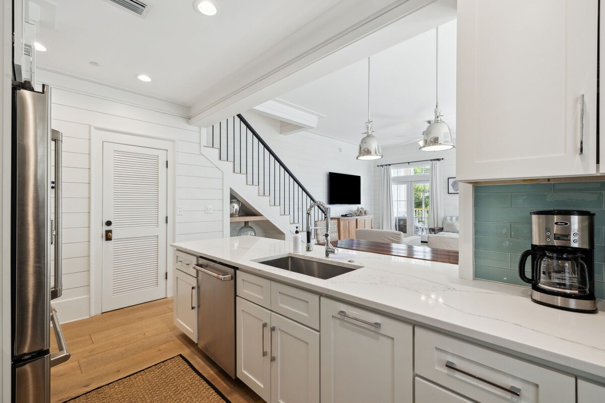A bright kitchen designed with modern features, highlighted by white cabinetry and sleek countertops. Stainless steel appliances are visible, accompanied by a coffee maker. An open-concept design allows for a view into the living area, where natural light fills the space.