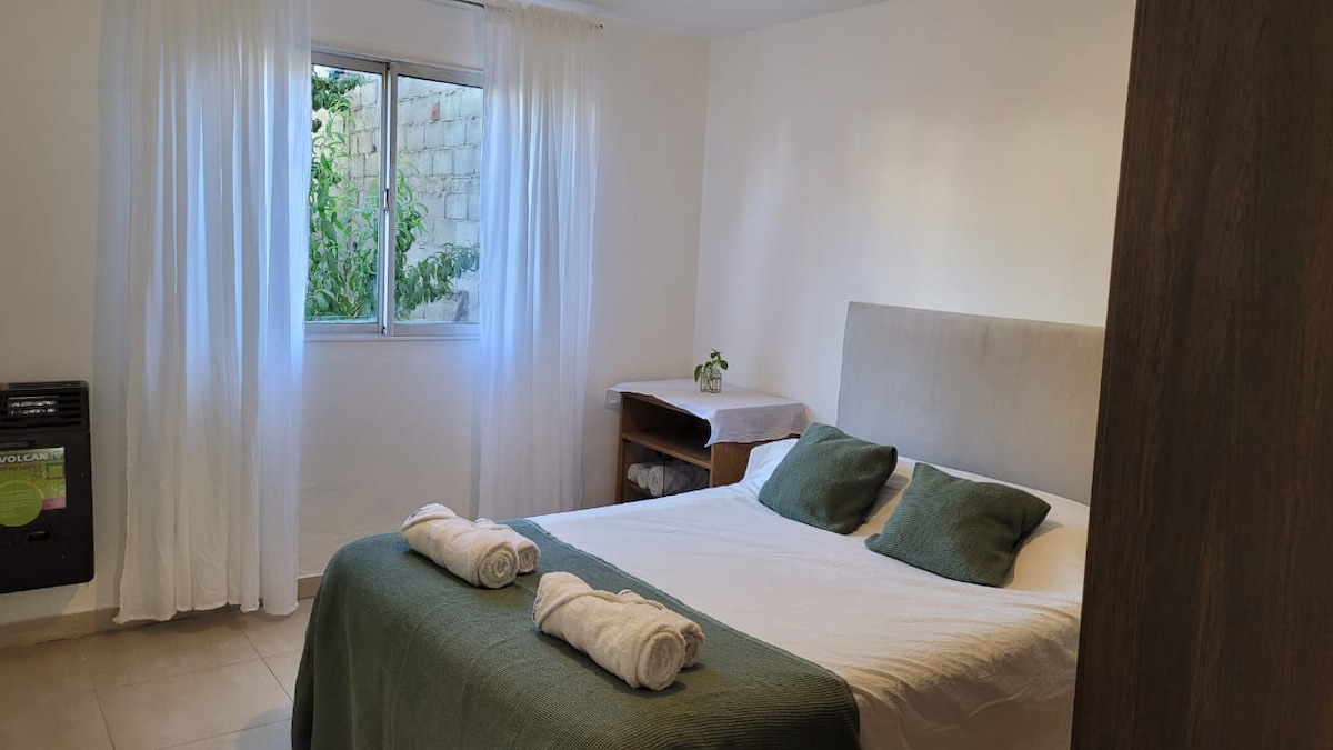A bedroom is presented with a spacious bed dressed in neutral linens and two green accent pillows. Fresh towels are neatly arranged at the foot of the bed. A window draped with sheer curtains allows natural light to brighten the room, complementing a wooden nightstand.