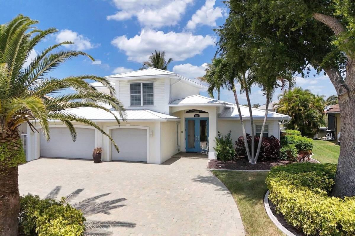 A charming exterior of a two-story villa is presented, featuring a light-colored facade and a prominent entrance with double blue doors. Lush landscaping surrounds the home, including palm trees and well-kept shrubs. A paved driveway leads to two garage doors.