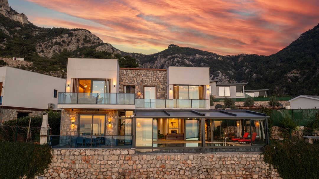 The villa showcases a contemporary design, featuring large glass windows and a stone facade. A spacious terrace overlooks the landscape, with comfortable seating options. The backdrop includes scenic mountains and a colorful sunset sky, enhancing the serene environment.