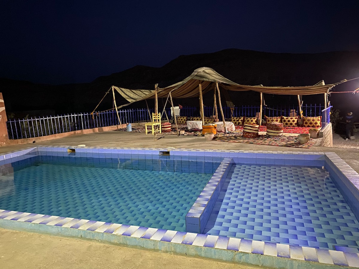 An inviting outdoor area is featured, showcasing a pool with a vibrant blue tiled surface. Adjacent to the pool, a traditional tent provides shaded seating with colorful cushions and intricately designed rugs. The surrounding landscape is softly illuminated under a night sky.