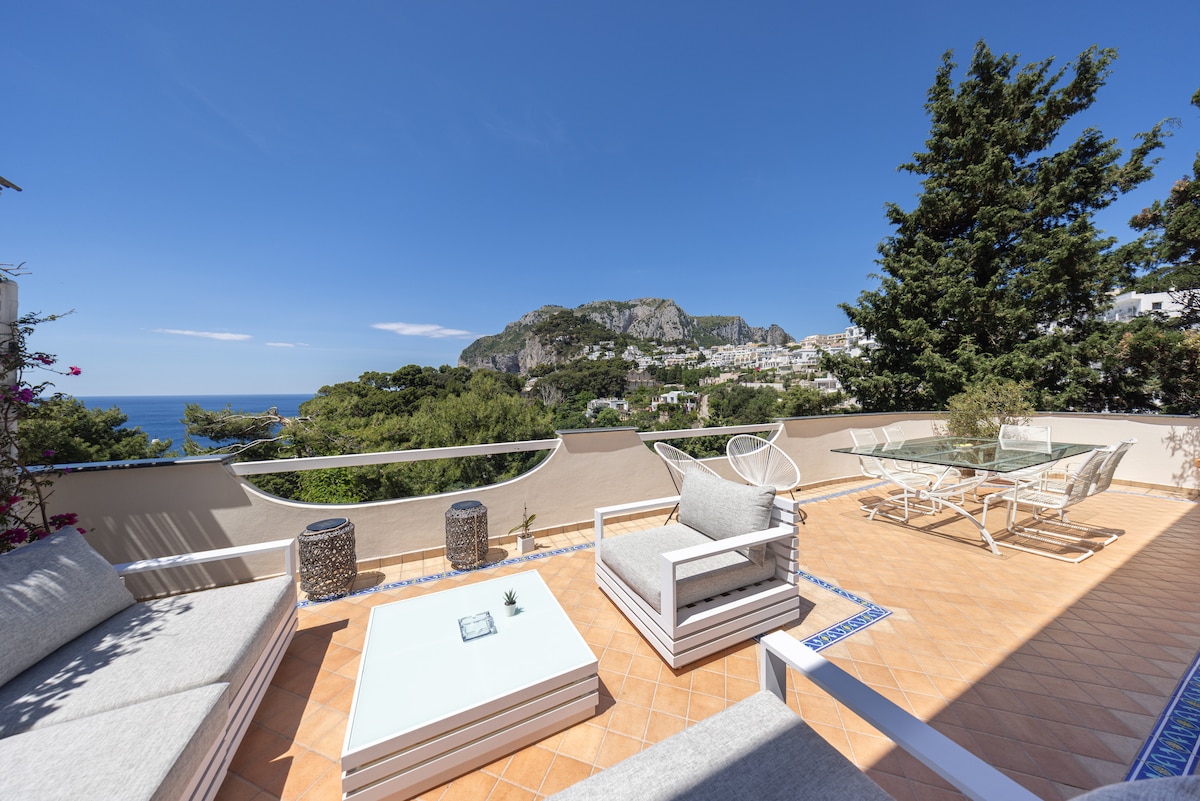 A spacious terrace features comfortable outdoor seating, including a soft grey sectional sofa and a square coffee table. Sunlight illuminates the area, offering clear views of the surrounding hills and the sea. Decorative elements include stone side tables, enhancing the outdoor dining experience.