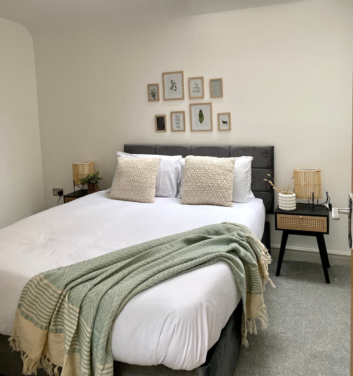A spacious bedroom features a King-sized bed with two decorative cushions. Artwork is arranged above the bed, while bedside tables with lamps are positioned on either side. A soft green throw drapes over the edge of the bed, adding a touch of texture to the neutral space.