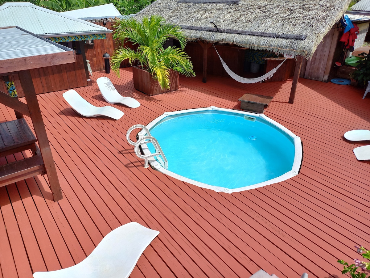 An inviting pool is surrounded by a spacious wooden deck, featuring sun loungers and a shaded seating area. A palm plant adds a tropical touch, while a hammock is visible, enhancing the relaxation space outdoors.