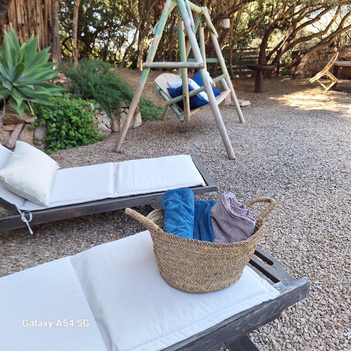 Two sun loungers with soft, neutral cushions are positioned on a gravel patio surrounded by greenery. A woven basket containing colorful towels rests on one of the loungers. In the background, a wooden swing hangs between trees, adding to the serene outdoor setting.