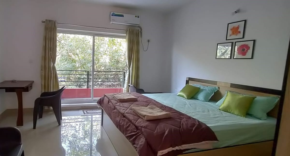 A spacious bedroom features a king-size bed adorned with light green and deep purple pillows. A window with sheer curtains opens to a view of greenery. A small table and chair are positioned near the window, complementing the room's minimalist decor.