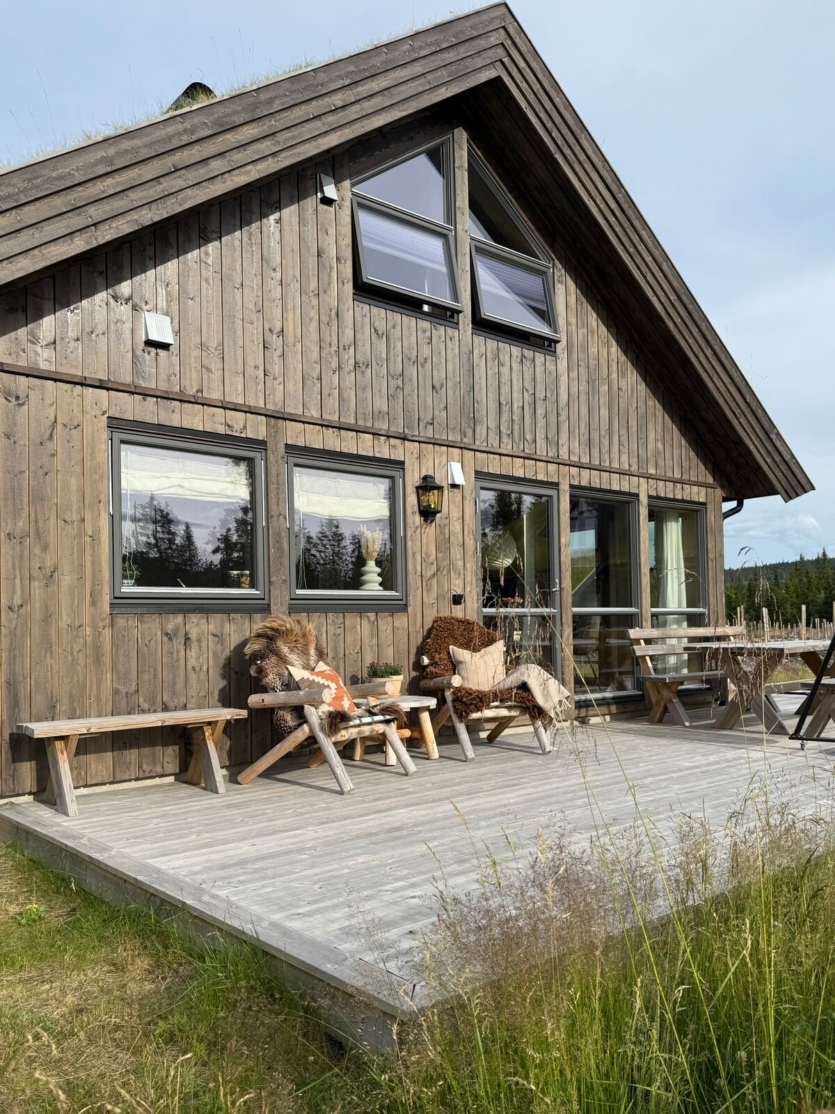 The exterior of the cabin showcases a wooden façade with a sloped roof. A spacious wooden deck is visible, furnished with several seating options. Two large windows allow natural light to enter, while surrounding tall grass adds to the serene outdoor environment.