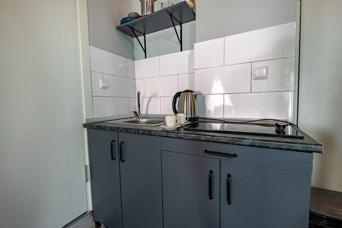 A compact kitchen area features a gray cabinet with sleek drawers and an integrated sink. A kettle is placed next to a white cup on the countertop, while light-colored tiles reflect a clean and modern aesthetic. Open shelving holds various kitchen items.