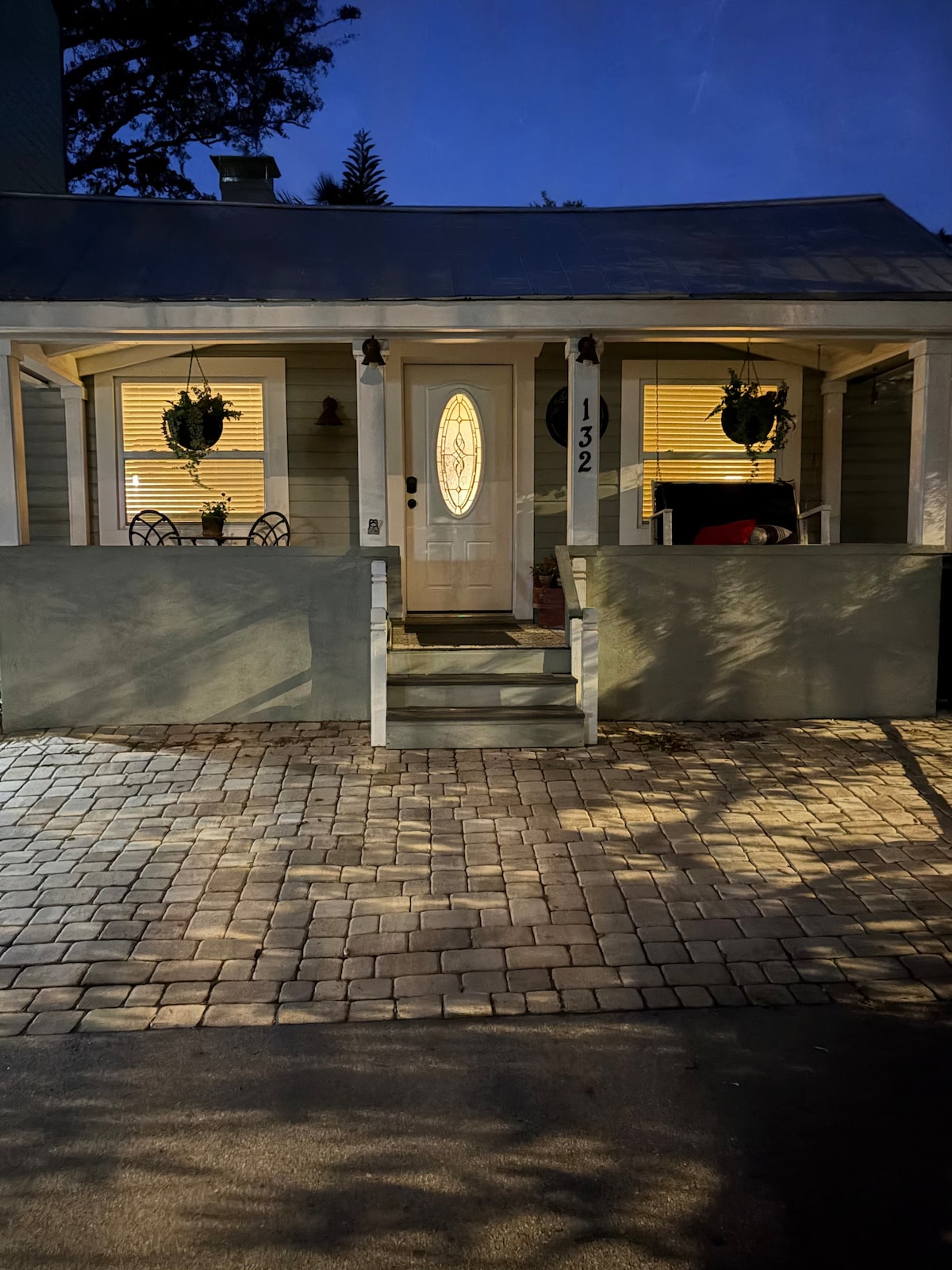 The inviting entrance of the historic cottage features a well-lit doorway framed by charming potted plants. A small outdoor seating area is visible, set with a table and chairs, while warm light spills from the interior windows, enhancing the welcoming ambiance.