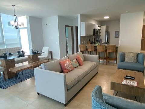 An open-concept living space is presented, featuring a comfortable light-colored sofa with decorative cushions. A wooden dining table accommodates several chairs, and a modern kitchen is visible in the background. Natural light fills the area through large windows, creating a welcoming environment.