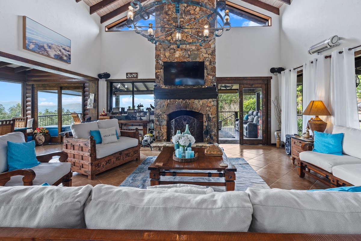 A spacious living area features high wooden ceilings and large windows that frame stunning views. A stone fireplace serves as a focal point, with comfortable seating arranged around a central wooden table, accented by decorative items and soft lighting.