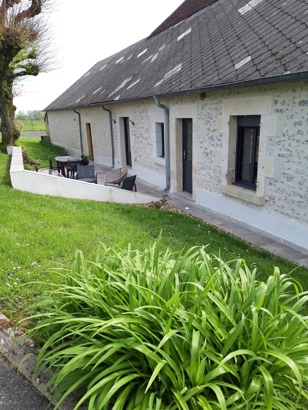 The exterior of the house features a blend of stone and plaster, with several large windows allowing natural light. A small terrace with outdoor seating overlooks a well-maintained garden, which includes lush green grass and flowering plants. A gentle slope leads from the grass to the terrace.
