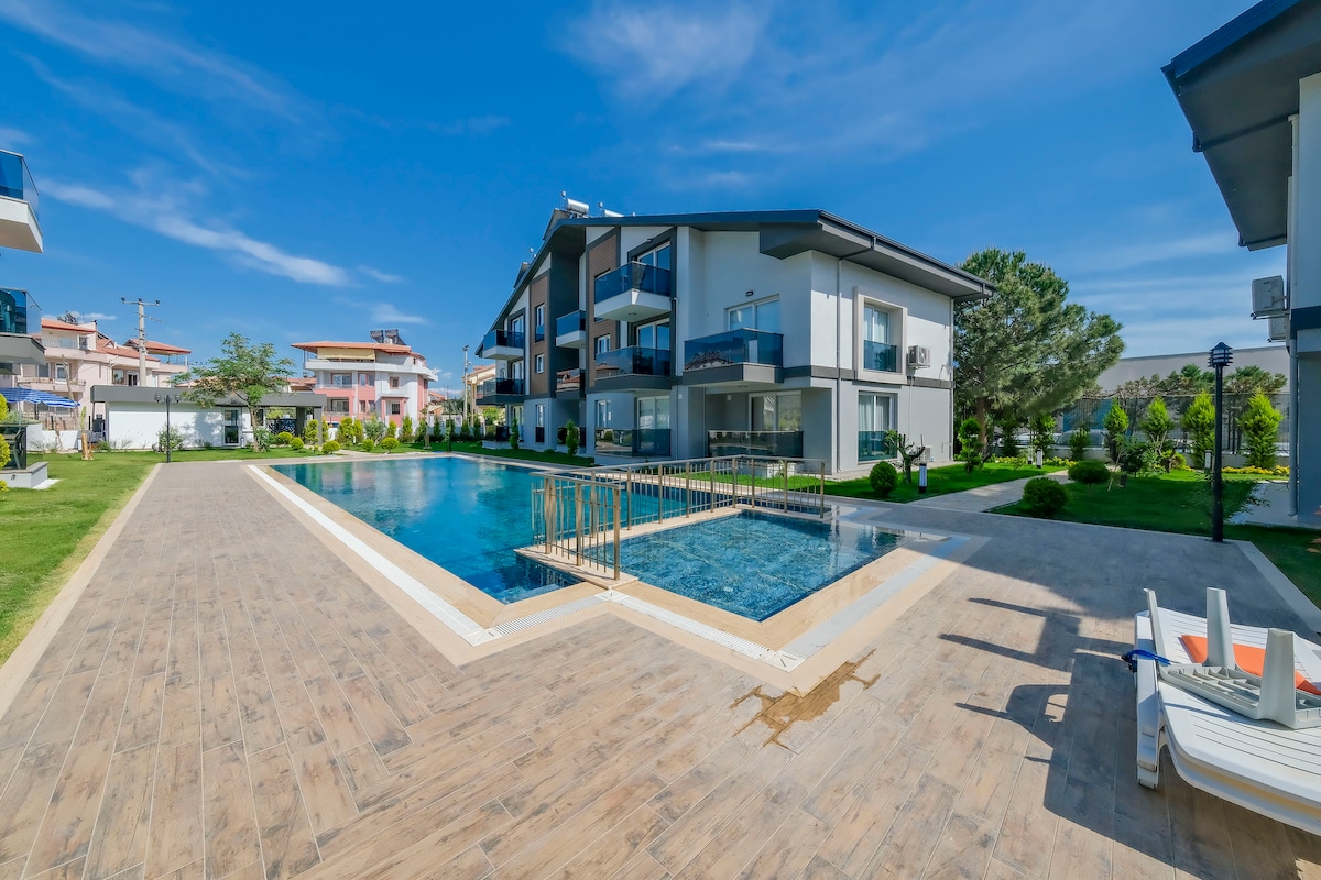 The image displays a well-maintained outdoor area featuring a large swimming pool surrounded by a paved deck. Lush greenery and plants are present, and several lounge chairs are positioned nearby. The two-story building provides a modern architectural backdrop.