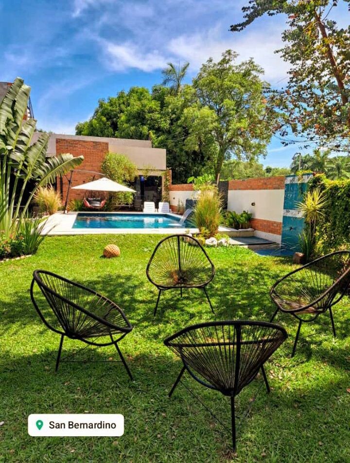 A spacious outdoor area features a well-maintained lawn surrounded by tropical plants. Five black chairs are arranged in a circle, facing a serene swimming pool with a sunshade nearby. Lush greenery provides a peaceful backdrop, enhancing the overall tranquil setting.