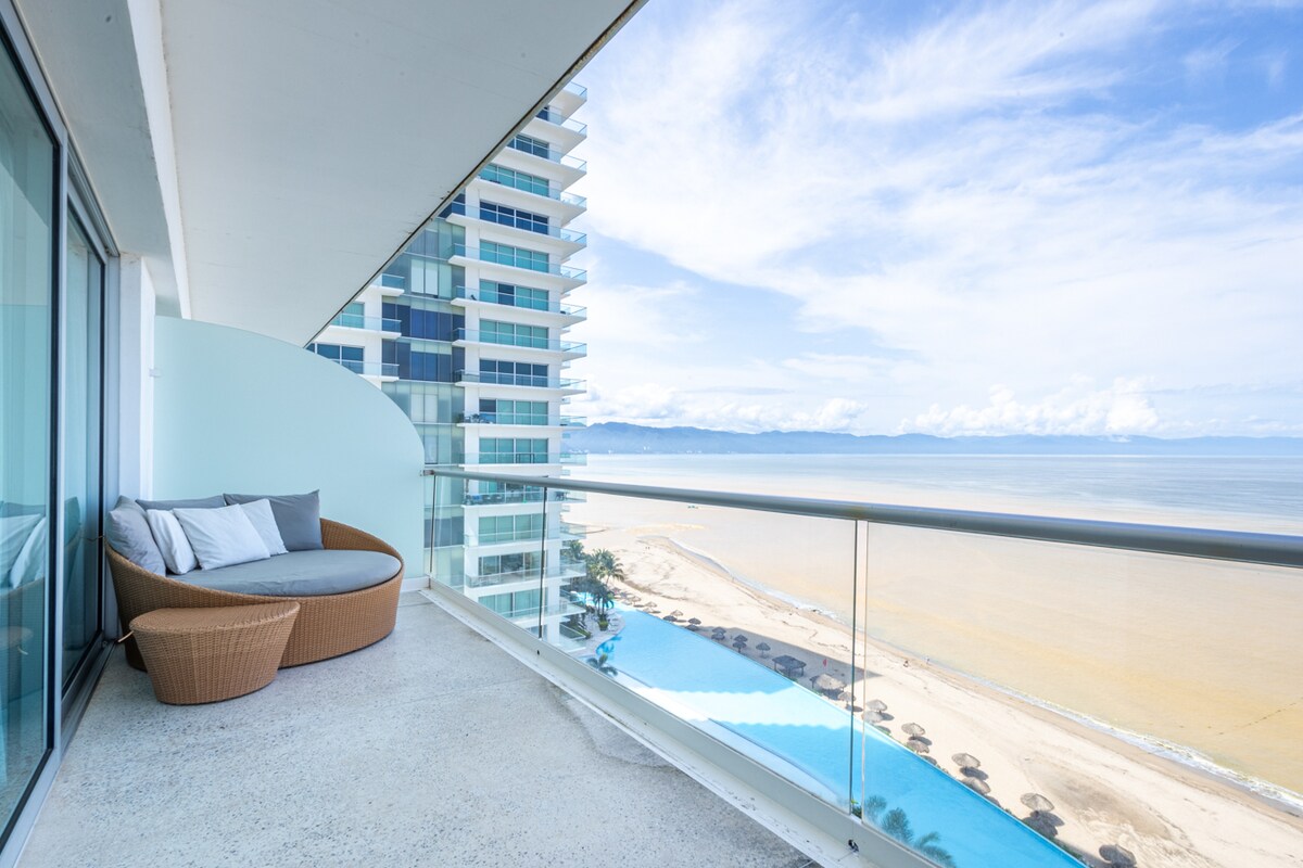 A spacious balcony is featured with a comfortable round seating area overlooking the beach and ocean. The panoramic view reveals a sandy shoreline and a serene pool area. Large glass doors provide access to the living space, enhancing the connection with the outdoors.