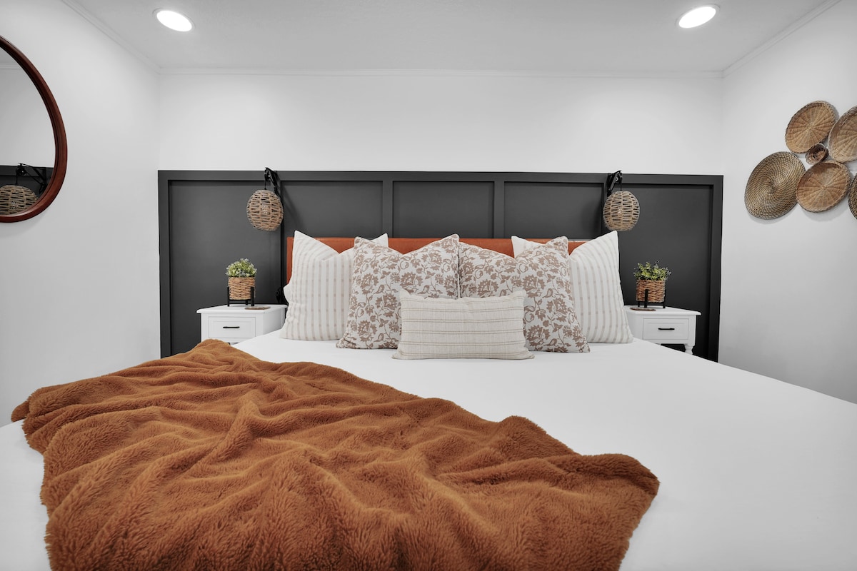 A serene bedroom features a spacious bed adorned with patterned pillows and a cozy brown throw blanket. Nightstands with small plants accentuate the sides, while decorative wall hangings add charm to the backdrop.