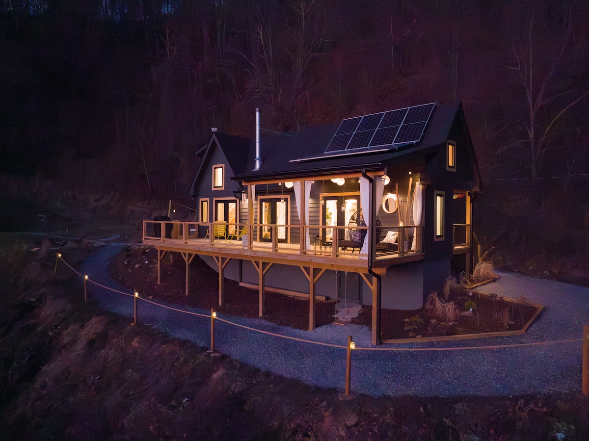 The exterior of the cabin is illuminated at dusk, showcasing modern architecture and eco-friendly solar panels. A spacious deck wraps around the front, with outdoor seating visible. The gravel driveway leads up to the cabin, enhancing the secluded mountain setting.