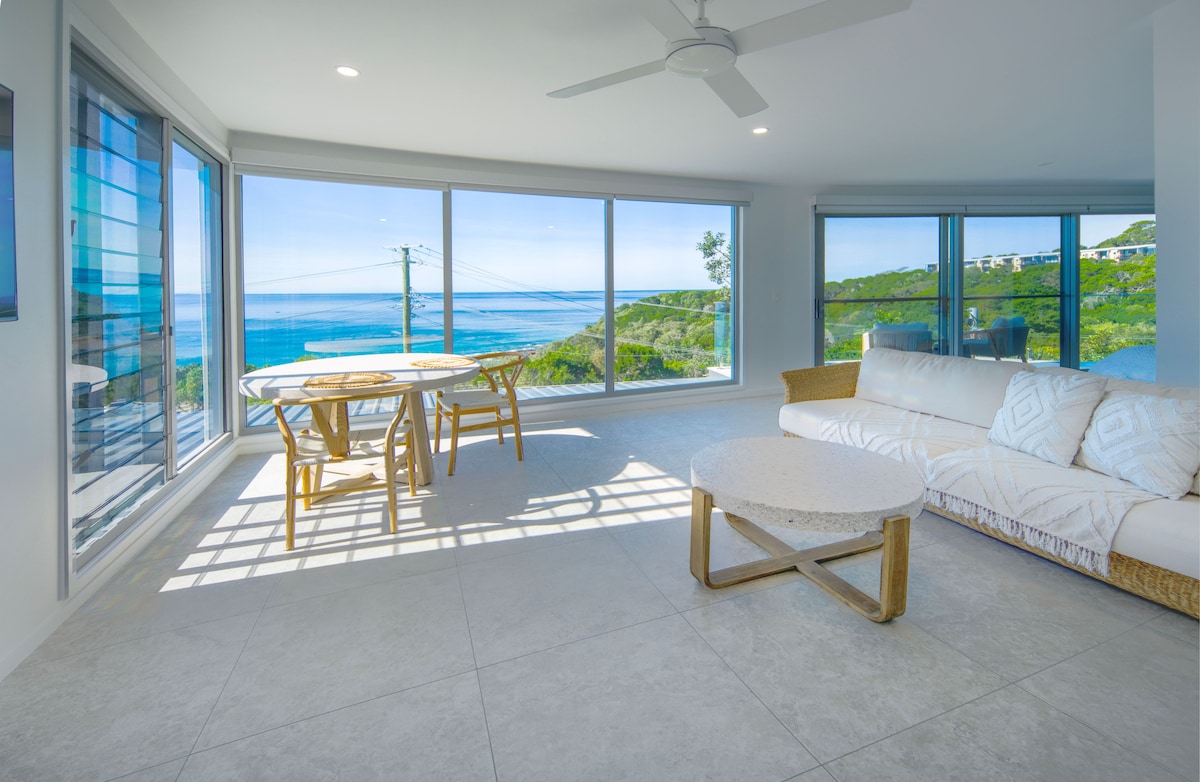 An open-plan living area features large floor-to-ceiling windows, allowing for natural light and showcasing panoramic ocean views. A light-coloured sofa and a round coffee table are positioned next to a dining table with four chairs, enhancing the spacious and airy atmosphere.
