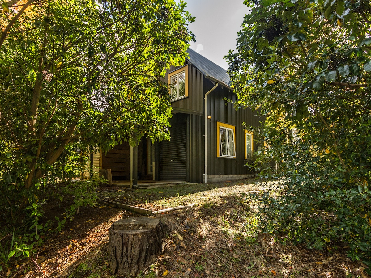 The exterior of the chalet is framed by lush greenery, creating a serene environment. The building features a dark exterior with bright yellow windows, enhancing its character. Sunlight filters through the trees, casting gentle shadows on the ground.