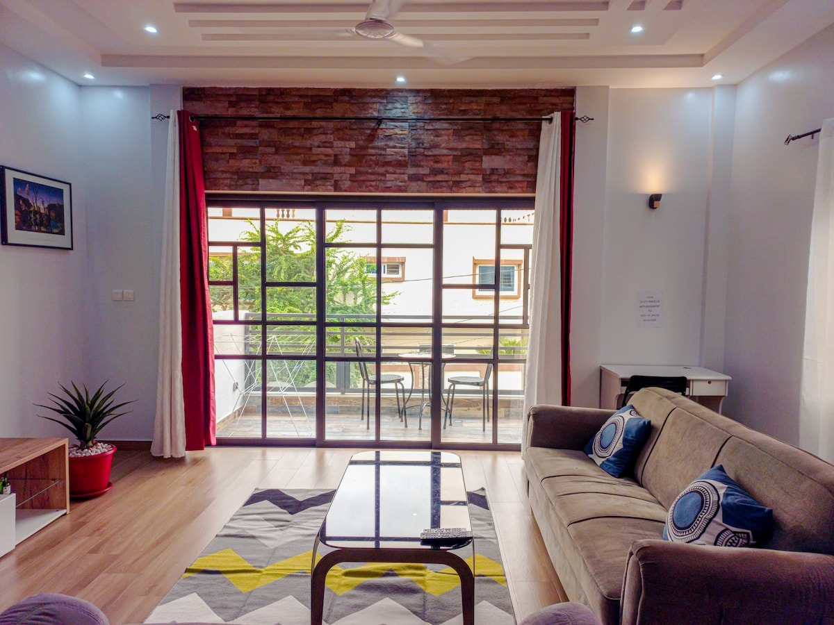 The living area features a comfortable sofa with decorative pillows, complemented by a coffee table. Large glass doors illuminate the space, providing a view of a balcony with outdoor seating. Indoor plants add a touch of greenery to the modern decor.