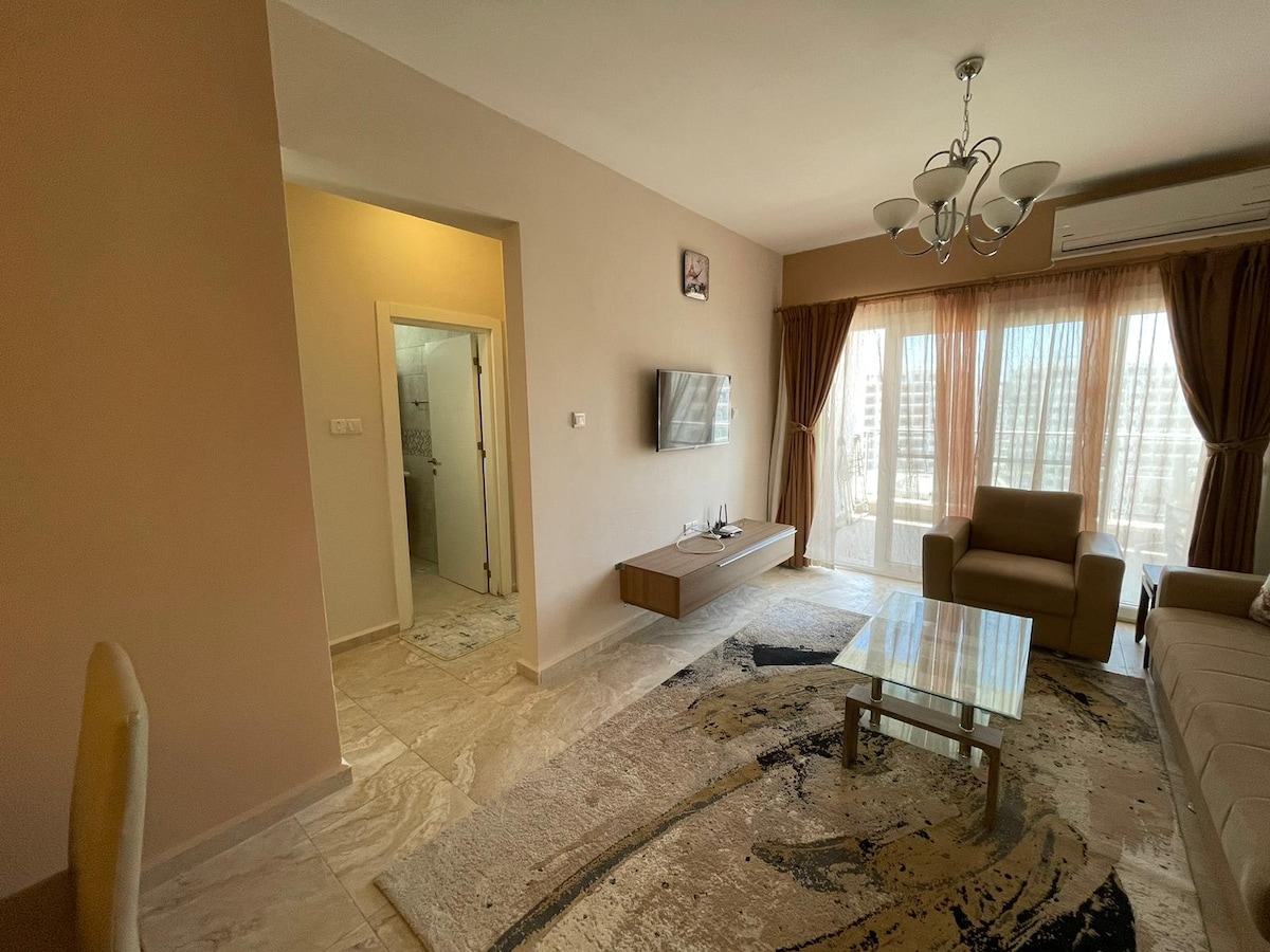 A spacious living area is highlighted by large windows that allow natural light, revealing a view of the surroundings. A comfortable sofa and armchair provide seating, while a glass coffee table sits atop a patterned rug. A flat-screen television is mounted on the wall.