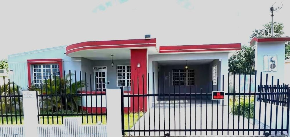 The exterior of a modern, one-story house is displayed, featuring a welcoming entrance with a red and gray color scheme. A gated fence surrounds the property, with lush greenery positioned next to the pathway. Ample space for parking is available under the carport.