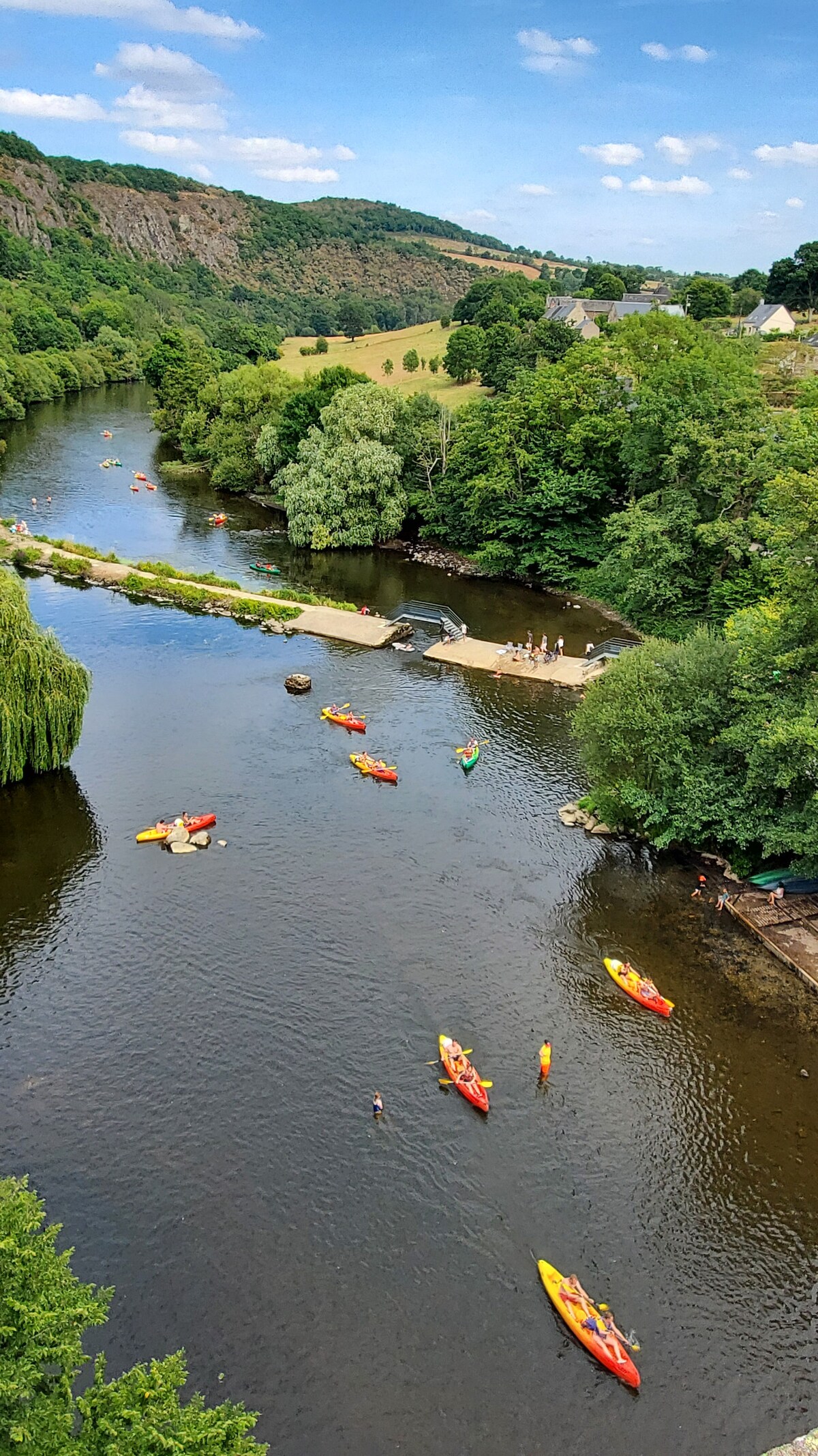 A scenic river scene is depicted, featuring several canoes in vibrant colors gliding across the calm water. Lush greenery lines the banks, with trees and grassy areas visible. A small sandy area along the river offers space for relaxation and activities.