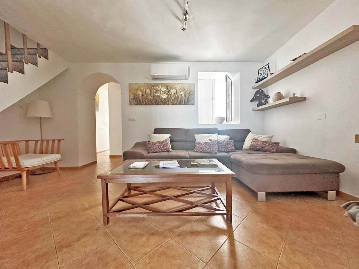 A spacious living area features a large L-shaped sofa adorned with decorative cushions. A wooden coffee table occupies the center, while an air conditioning unit is mounted above. Light enters through a window beside the stairway, illuminating the warm tile flooring.