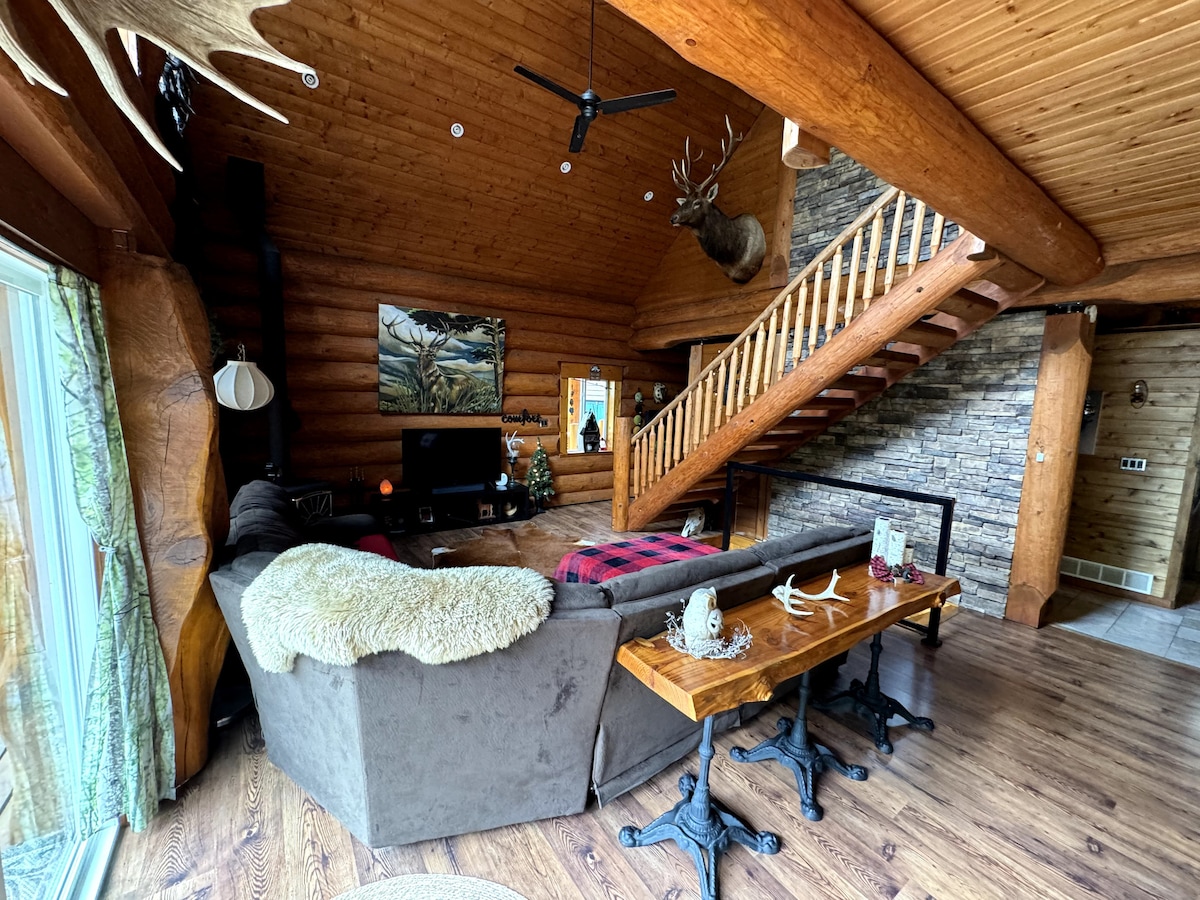 A spacious living area is highlighted by high wooden ceilings and natural light from large windows. A comfortable sectional sofa is adorned with plush throws, facing a mounted TV. A rustic wooden table complements the decor, while a staircase leads to the upper level.