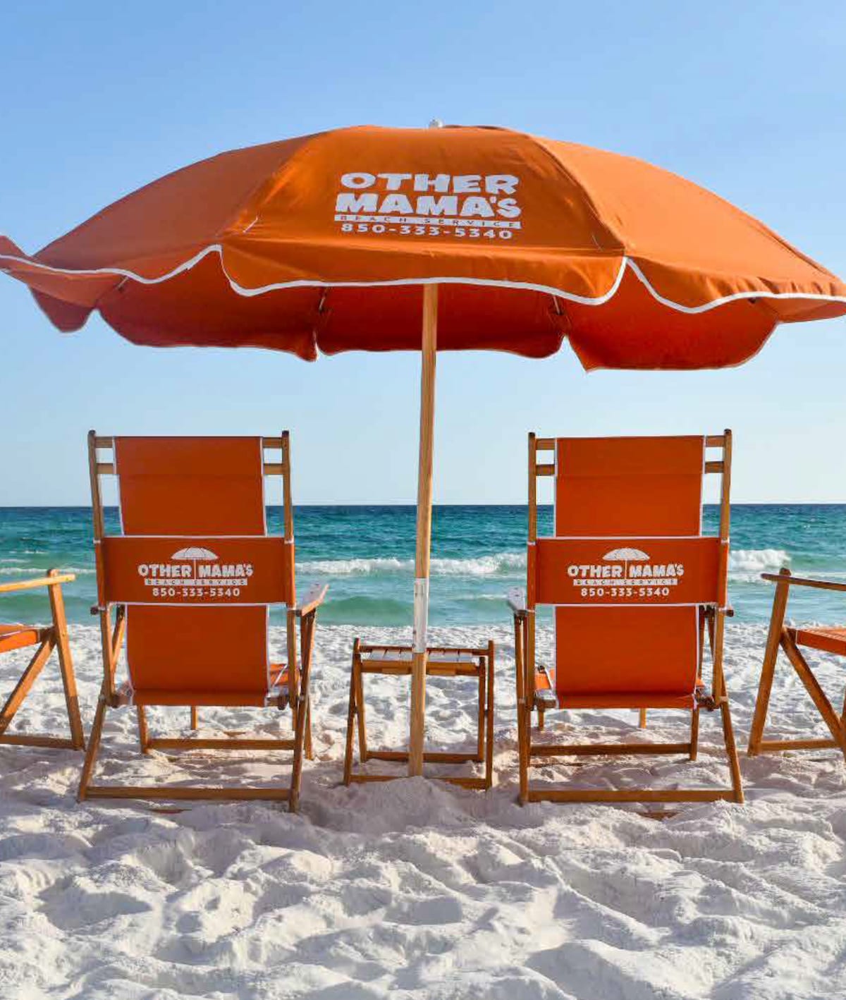 Two wooden beach chairs are situated under a large orange umbrella, labeled with 'OTHER MAMAS.' The chairs face the calm turquoise waters of the ocean, with soft white sand surrounding them, creating a relaxing beach setup.