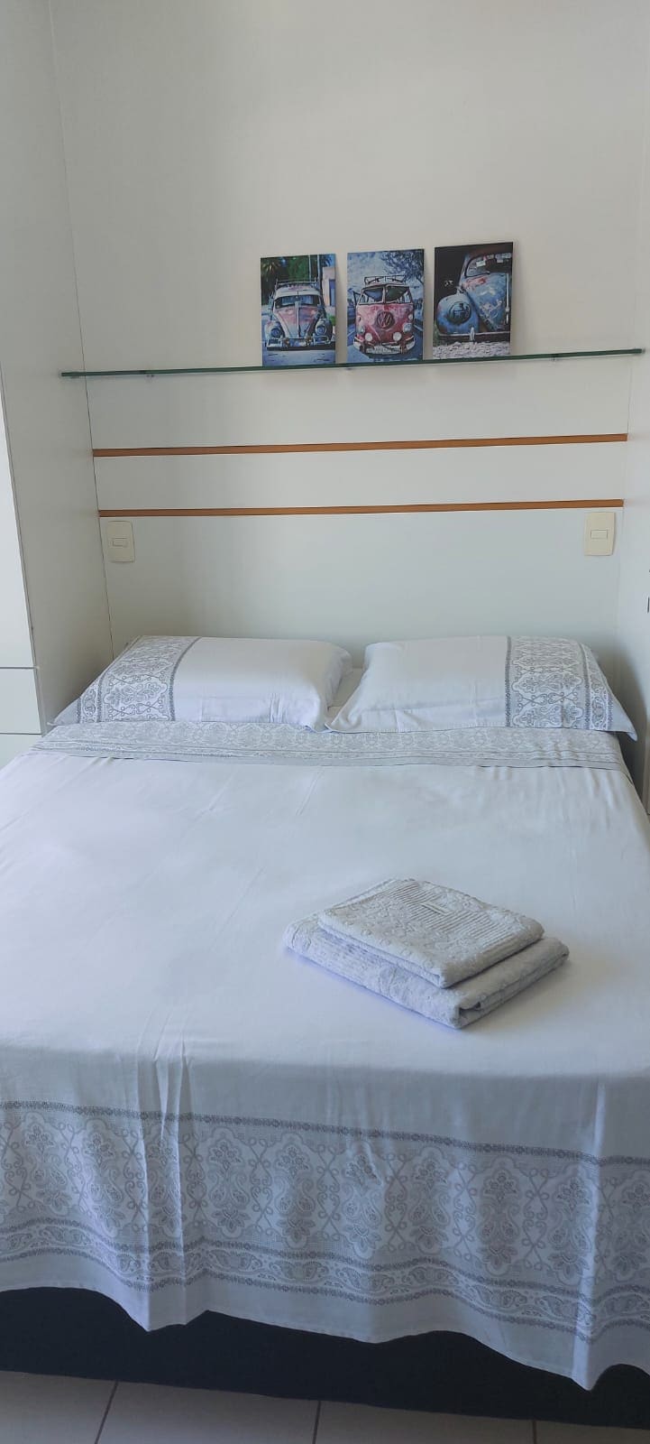 A cozy sleeping area features a spacious bed with crisp white linens, neatly arranged with folded towels. Above the bed, three framed art pieces add a touch of character to the minimalist design.