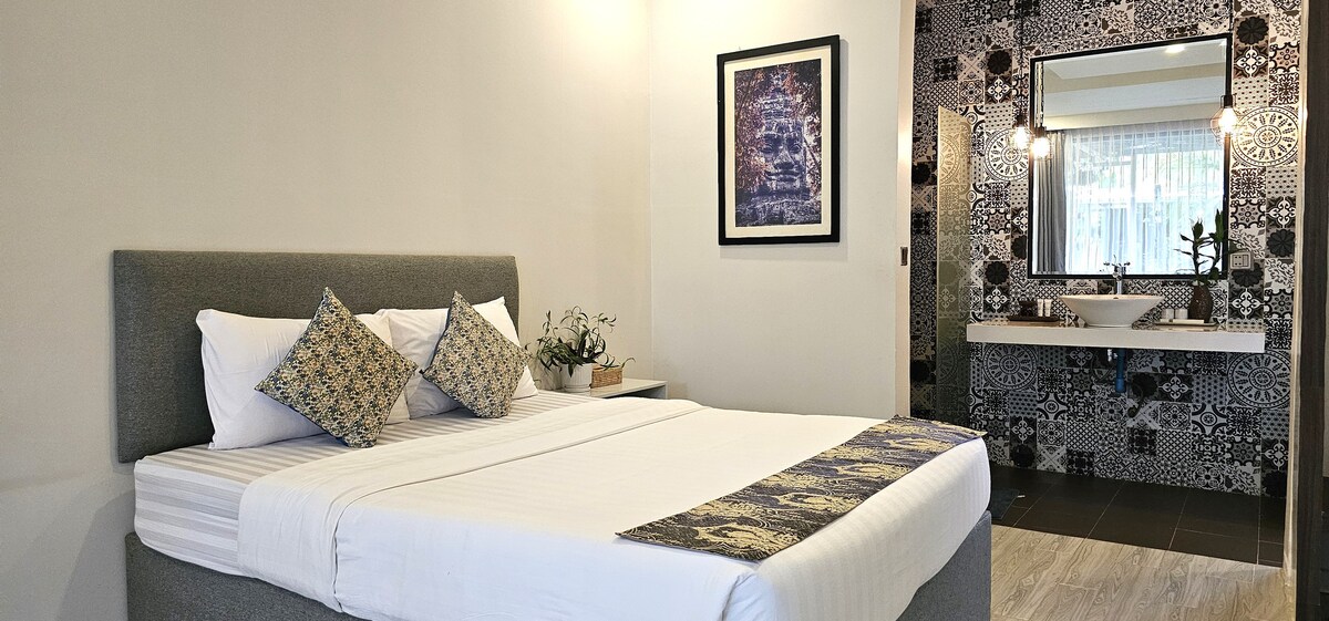 A well-appointed room features a spacious bed with decorative pillows, complemented by a minimalist design. A large mirror and sink area are visible in the background, alongside patterned tiles that add a unique touch to the space.