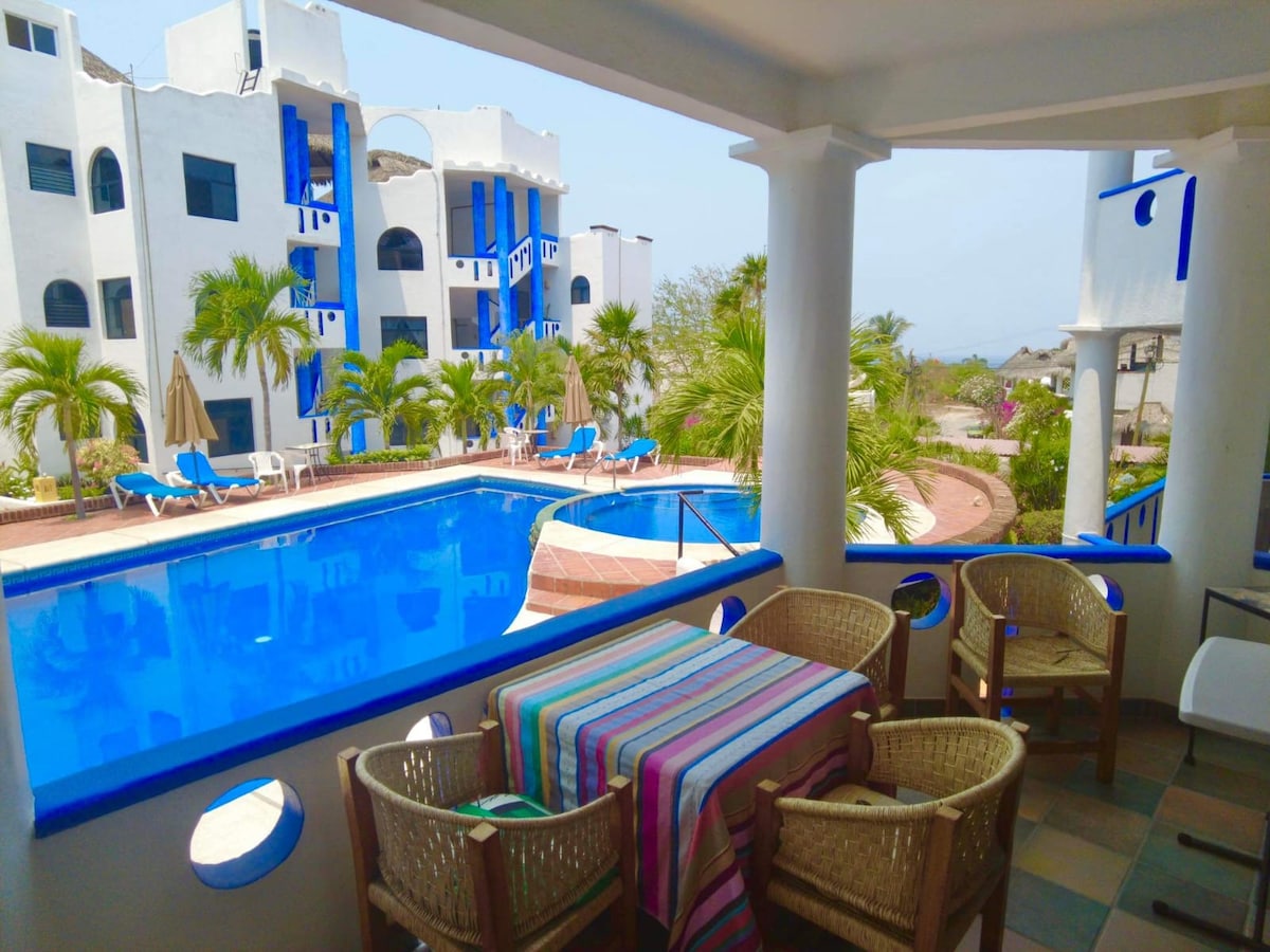A private terrace offers a view of the main pool surrounded by lush landscaping. The area features a table with striped tablecloth and four wicker chairs, while lounge chairs and umbrellas are positioned near the pool for relaxation.
