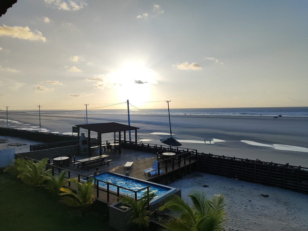 The image captures a serene beachfront scene during sunset, with a pool visible in the foreground. A wooden deck with seating areas overlooks the expansive sandy beach, framed by palm leaves. The horizon is illuminated by the sun setting over the ocean, creating a peaceful atmosphere.