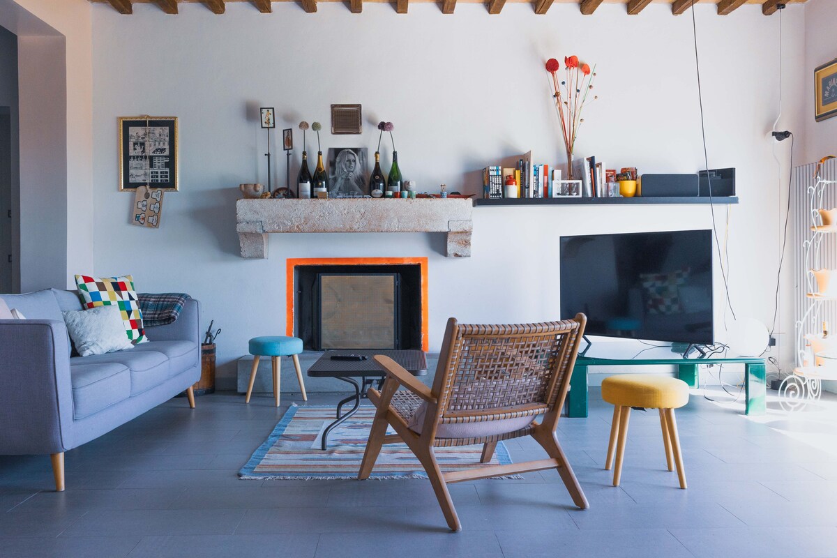 A spacious living area features a stone fireplace surrounded by decorative items. A gray sofa and a wooden chair provide seating, with a coffee table positioned at the center. A wall-mounted television and a shelf filled with books complete the modern yet cozy atmosphere.