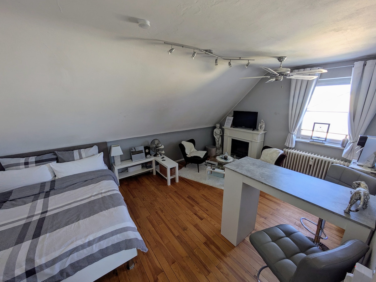 A modernly furnished room features a spacious bed with layered bedding, wooden flooring, and a large window providing natural light. A seating area includes two chairs and a small table, while a desk with a chair is positioned near the window. A TV is visible on a stand.