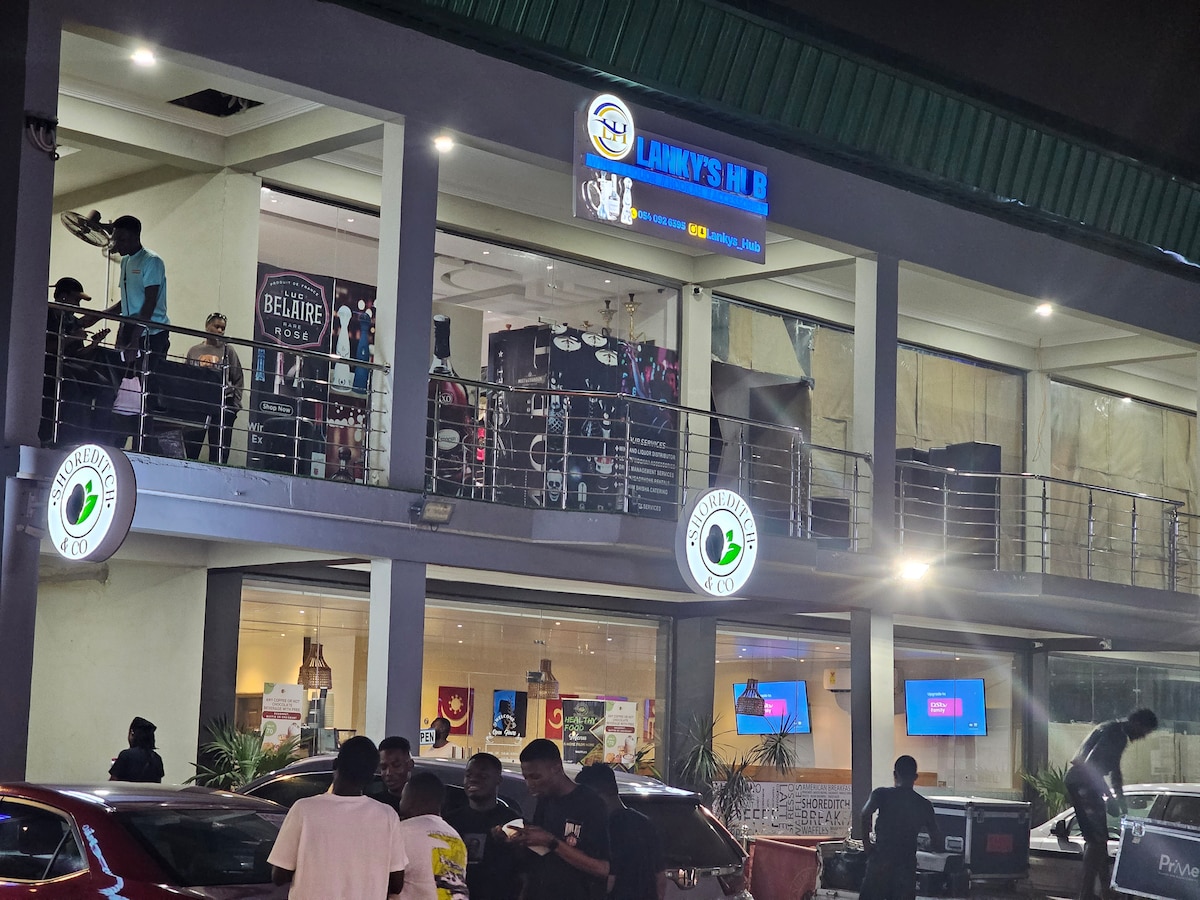 A lively scene is depicted outside a multi-level venue at night. The building features a modern design with large glass windows and illuminated signage. Groups of people are gathered in front, while colorful lights enhance the atmosphere. Vehicles are parked nearby.