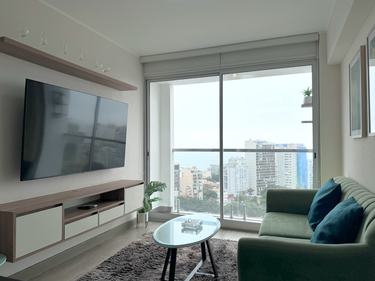 A contemporary living space is presented, featuring a large flat-screen television mounted on a wooden panel. A glass coffee table is positioned in front of a stylish green couch. Floor-to-ceiling windows provide striking views of the city and ocean.