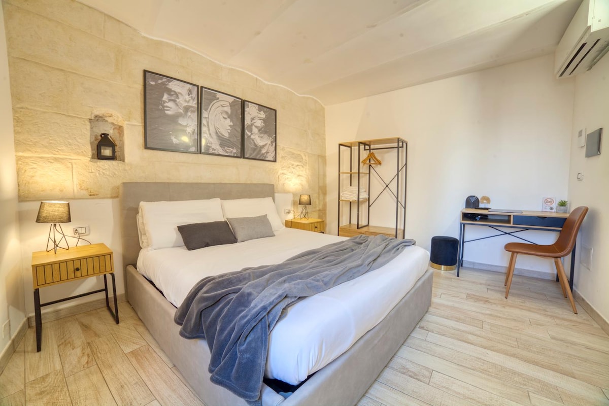 A cozy bedroom features a spacious bed dressed in crisp white linens, accented by a soft gray throw. Decorative wall art complements the light-colored stone walls. A wooden desk and chair provide a workspace, while bedside lamps offer gentle illumination.