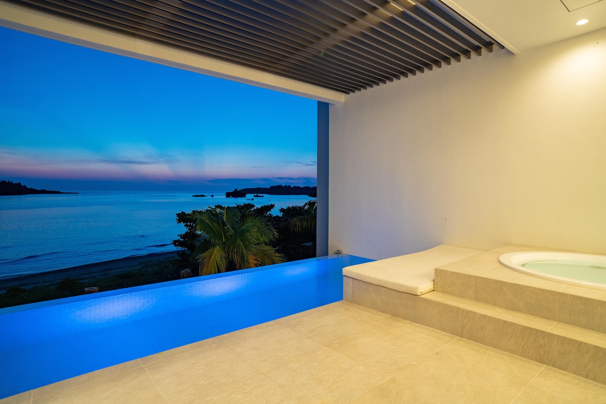 An inviting area features a private pool with a stunning view of the ocean at dusk. The smooth, cool tiles set a calming ambiance, while the gently lit whirlpool stands beside the pool. Large windows enhance the openness and connection with the serene outdoor landscape.
